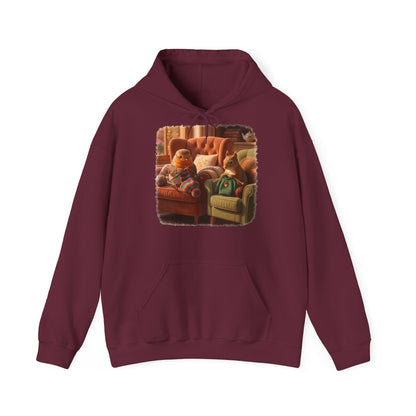 Ein dunkelroter Kapuzenpullover mit einem quadratischen Grafikdruck auf der Brust, der eine gemütliche Wohnzimmerszene zeigt: Ein Rotkehlchen sitzt neben einem Eichhörnchen im grünen Pullover auf einem Sessel, und beide stricken.