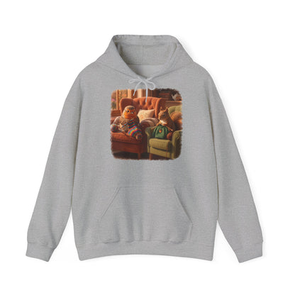 Ein hellgrauer Kapuzenpullover mit einem quadratischen Grafikdruck auf der Brust, der eine gemütliche Wohnzimmerszene zeigt: Ein Rotkehlchen sitzt neben einem Eichhörnchen im grünen Pullover auf einem Sessel, und beide stricken.