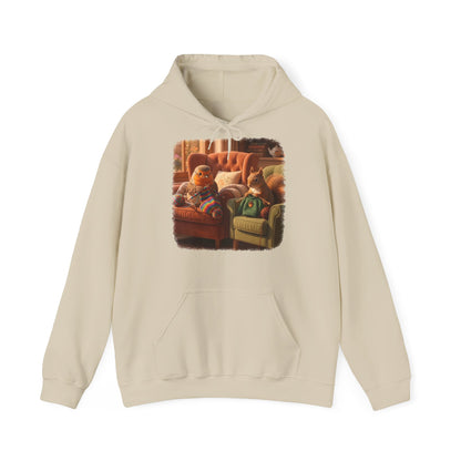Ein beiger Kapuzenpullover mit einem quadratischen Grafikdruck auf der Brust, der eine gemütliche Wohnzimmerszene zeigt: Ein Rotkehlchen sitzt neben einem Eichhörnchen im grünen Pullover auf einem Sessel, und beide stricken.